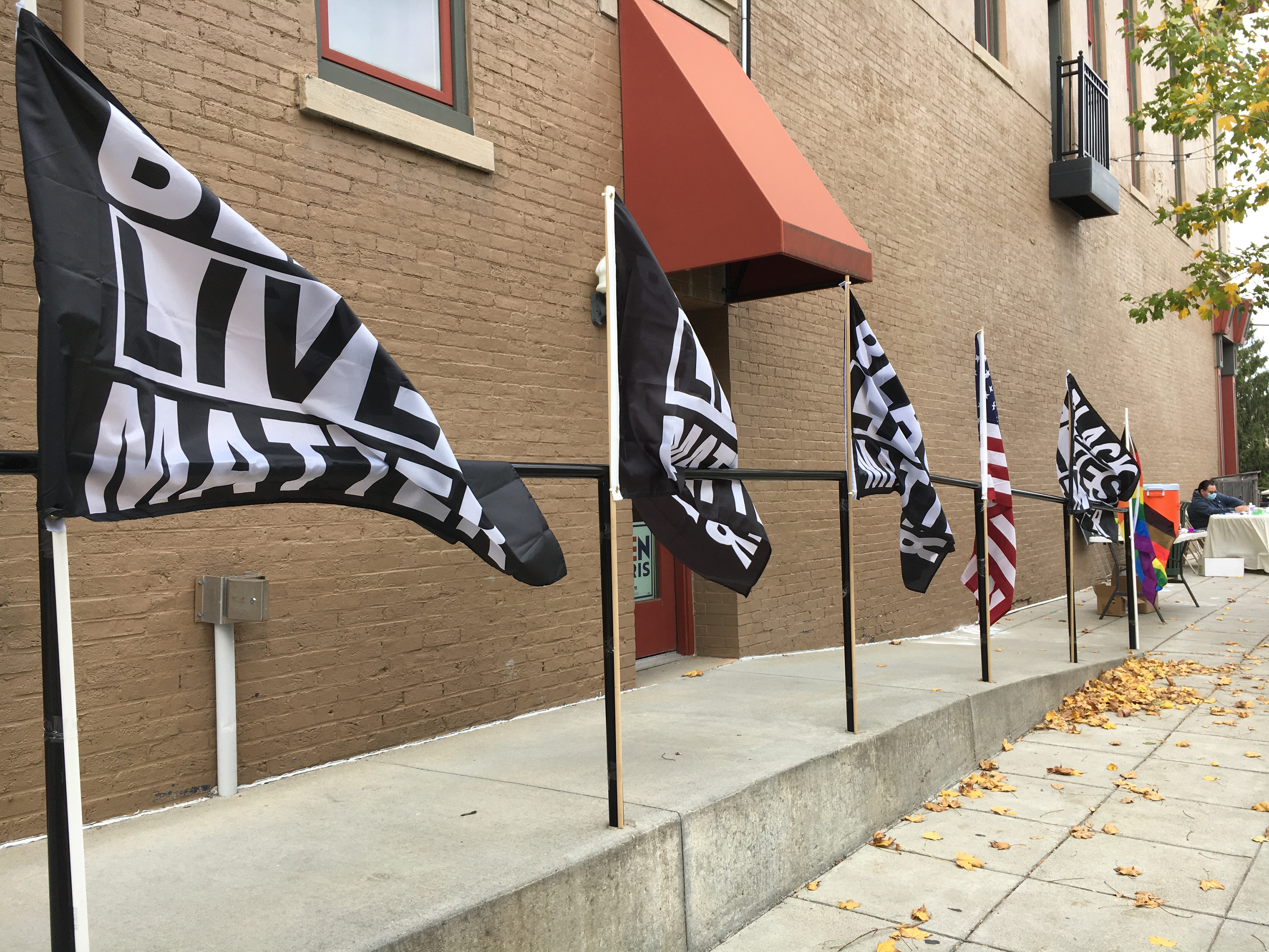 Four Black Lives Matter flags, an American Flag, and a gay pride flag are set up on the sidewalk railings in front of a brick building.