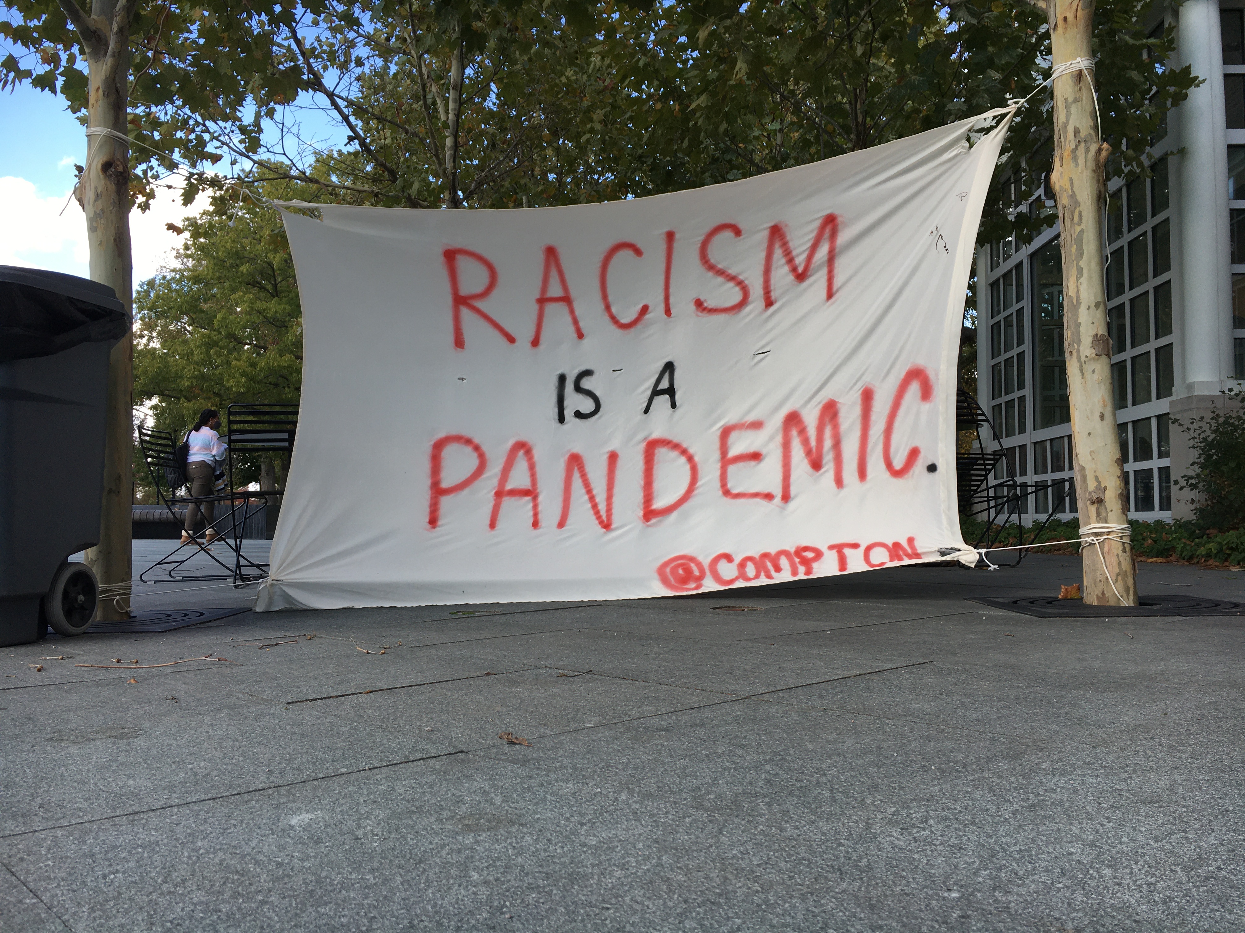 A canvas sign hung between two trees reads “Racism is a pandemic,” @compton.