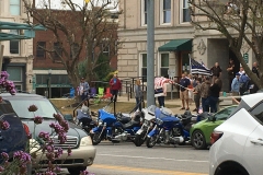 Beyond a street with some motorcycles and cars is a crowd of people with signs like “we support the police” and blue-striped American flags on the courthouse lawn.