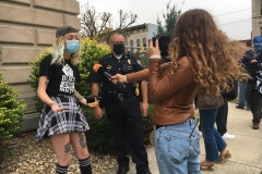 A journalist with a microphone and a camera interviews one of the protest organizers from Putnam County Greater Good while a police officer stands off to the side.