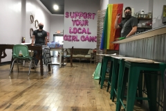 Interior of a shop with Black Lives Matter signs and poster supplies. A gay pride flag hangs on wall. Another has balloons that say, “support your local girl gang.” Two people are visible at the poster-making tables.