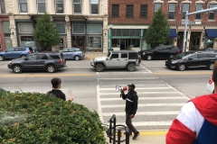 A black man speaks into a red and white bullhorn from the sidewalk in front of the grass embankment the protesters are on.