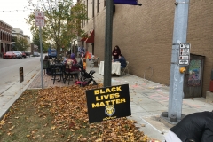Behind a DePauw University Black Lives Matter sign, a few families and individuals are gathered around tables along the sidewalk.