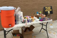 A table with a water cooler, paints, chalk, paint brushes, templates, and more art supplies is set up on the sidewalk.