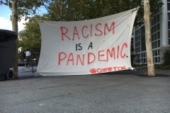 A canvas sign hung between two trees reads “Racism is a pandemic,” @compton.